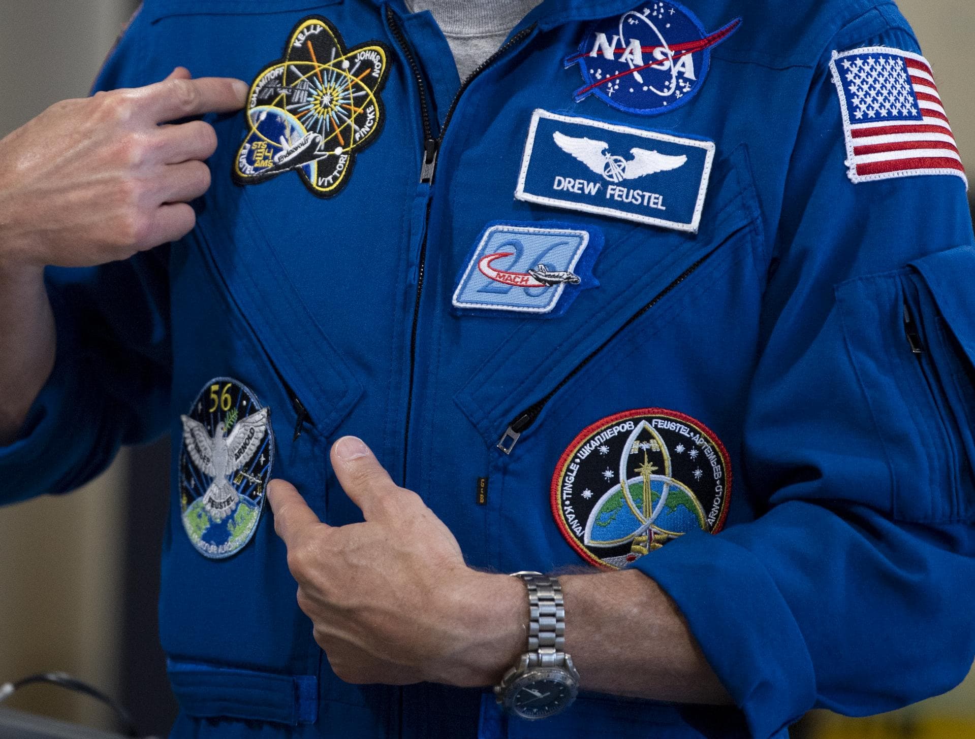 NASA astronaut Drew Feustel pointing to the mission patches stitched onto his blue flight suit.